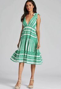 Grön- och vitmönstrad klänning med djup V-ringning, utställd kjol och knytbara axelband, tillverkad av lätt tyg.