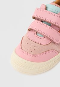 Ροζ δερμάτινο και σουέντ αθλητικό παπούτσι με δύο λουριά βέλκρο, με γλώσσα σε ανοιχτό μπλε και διακοσμητικές οπές στη μύτη. Κρέμ λαστιχένια σόλα.