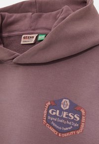 Felpa grigio-viola realizzata in tessuto morbido con un grande logo circolare che presenta la scritta "GUESS" in bianco e rosso. Etichetta ecologica visibile.