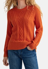 Pull en maille orange avec motifs texturés, détails en torsades et poignets et ourlet côtelés. Porté avec un jean bleu.
