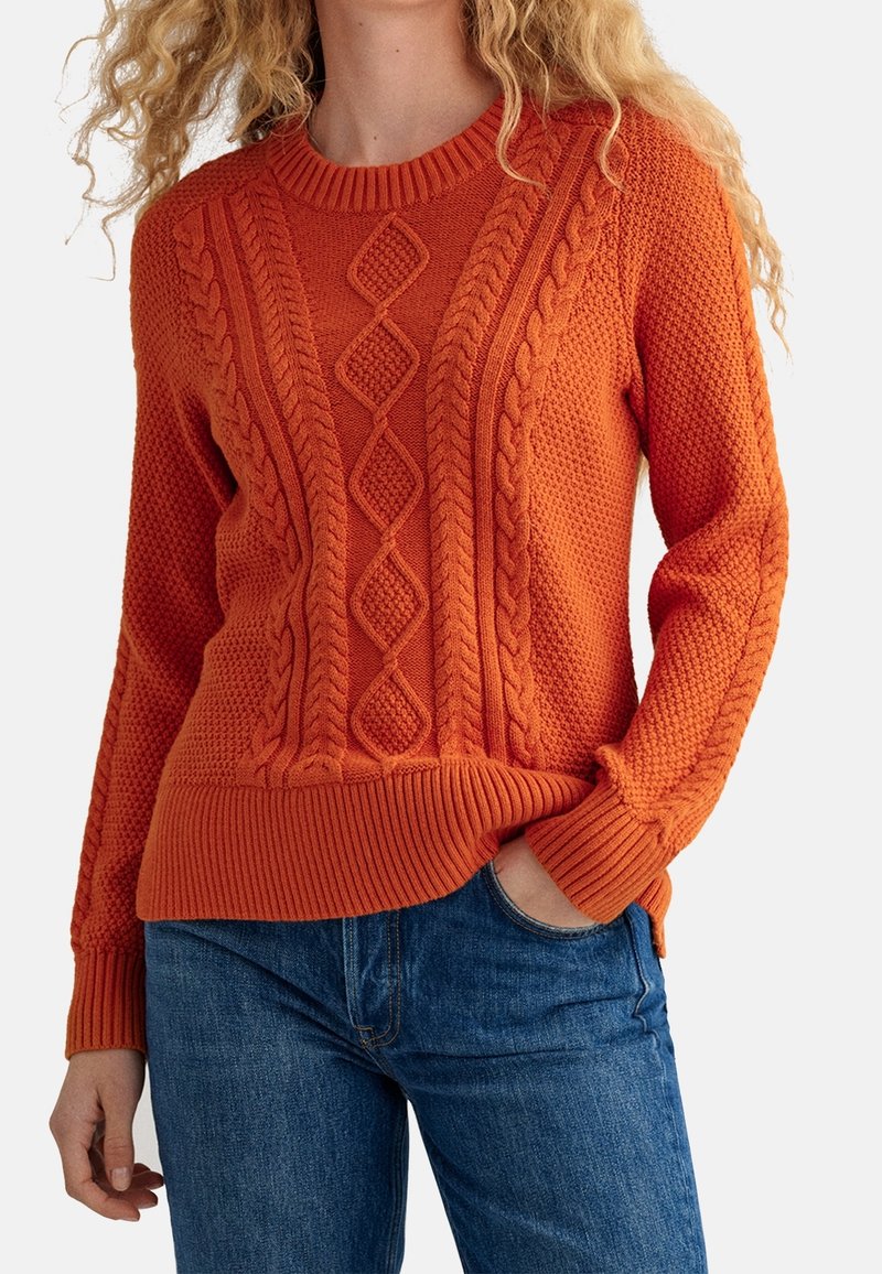 Pull en maille orange avec motifs texturés, détails en torsades et poignets et ourlet côtelés. Porté avec un jean bleu.