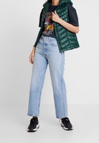 Osoba ubrana w jasnoniebieskie jeansy o wysokim stanie, czarną koszulkę z grafiką, zieloną pikowaną kurtkę bez rękawów i czarne sneakersy, stojąca na białym tle.