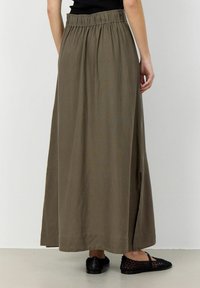 Gonna maxi in tessuto verde oliva con una cintura elastica arricciata, dotata di uno spacco laterale per facilitare i movimenti. Abbinata a ballerine nere intrecciate.