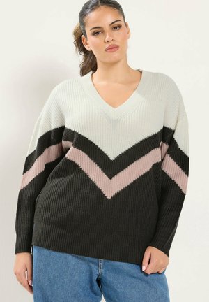 Femme portant un pull tricoté à motif chevron blanc, noir et rose avec col en V et un jean bleu, debout devant un fond uni.