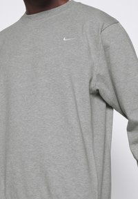 Sudadera gris claro hecha de suave algodón, con cuello redondo, mangas largas y un pequeño logo blanco de Nike en el pecho.