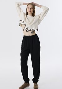 Sudadera crema cortada con "Muse" en script negro, combinada con pantalones negros holgados que cuentan con cinturilla elástica y bolsillos laterales.
