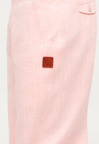 Svjetloružičaste hlače s teksturom, bočne džepove i malenim smeđim kvadratnim oznakom marke blizu džepa.