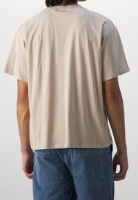 Beige kortærmet t-shirt lavet af blød bomuld, med en afslappet pasform, rund halsudskæring og minimale syninger. Set fra bagsiden.