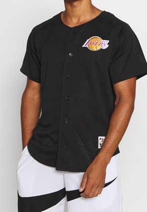 Mężczyzna ubrany w czarną koszulę na guziki drużyny Los Angeles Lakers oraz białe spodenki z czarnym wzorem, stojący z jedną ręką w kieszeni.