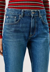 Jeans de mezclilla azul oscuro con un diseño clásico de cinco bolsillos, costuras claras y un cierre de botón en la cintura. La silueta es ajustada.