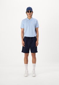 Polo shirt bleu clair, shorts navy, baskets blanches, chaussettes blanches, casquette navy, lunettes de soleil noires ; tenue d'été décontractée avec une coupe ample.
