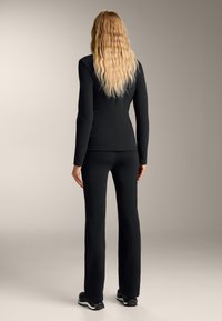 Czarna dopasowana bluzka z długim rękawem i pasujące spodnie, wykonane z gładkiego materiału o eleganckim fasonie. Modelka ma falowane blond włosy i nosi czarne sneakersy.