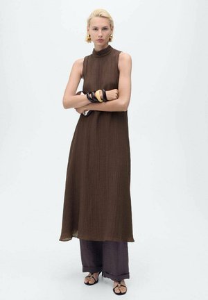 Langes braunes ärmelloses Kleid mit hohem Kragen und strukturiertem Stoff, das über dunkelvioletten weit geschnittenen Hosen getragen wird, ergänzt durch schwarze Sandalen.