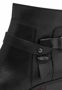 Botín negro de piel con un diseño elegante, una correa con hebilla y un acabado texturizado, con un tacón de madera oscura ligeramente elevado.