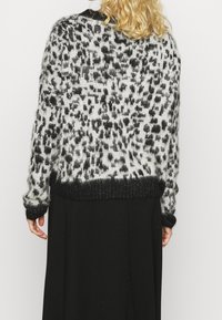 Eine blonde Person, die einen flauschigen Pullover mit Leopardenmuster und schwarzen Bündchen und Saum trägt, kombiniert mit einem langen schwarzen Rock, von hinten gezeigt.