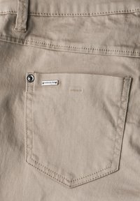 Ljusbeige tyg med en texturerad yta, med en bakficka med en silverknapp och en liten metalldetalj nära övre kanten.
