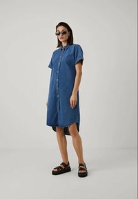 Φόρεμα από τζιν, κοντά μανίκια, με κουμπιά μπροστά, μήκος μέχρι το γόνατο, ανοιχτό μπλε χρώμα, με ίσια γραμμή και πλαϊνές σκισίματα. Φορεμένο με σκούρες σανδάλια.