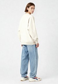 Jeune homme aux cheveux mi-longs portant un sweat-shirt crème surdimensionné, un jean bleu clair ample, et des baskets blanches avec des détails rouges et bleus.