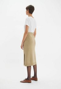 Jupe midi en tricot beige avec une ceinture côtelée et une fente au dos, associée à un t-shirt ajusté blanc et des chaussures marron. Détail de tissu texturé visible.