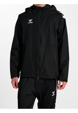 Mężczyzna ubrany w czarną bluzę z kapturem na zamek oraz czarne spodnie z białymi logo Hummel na piersi i udzie, stojący na białym tle.