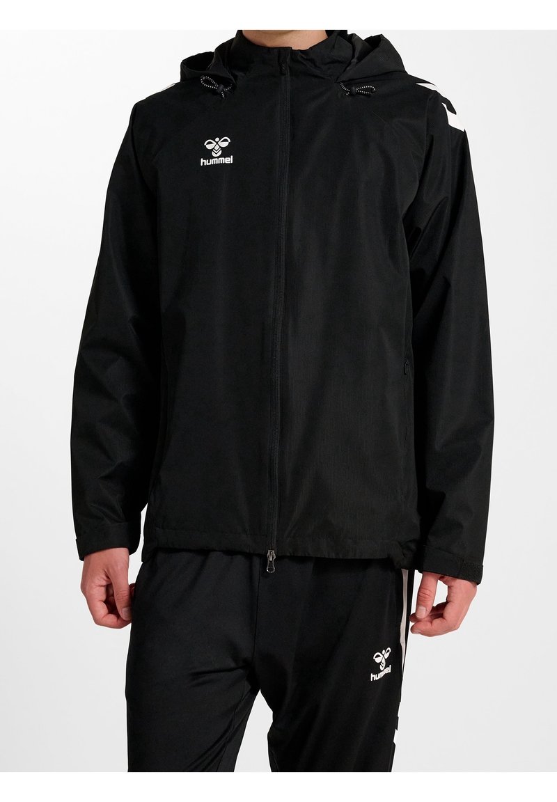 Homme portant une veste à capuche noire zippée et un pantalon noir avec des logos blancs Hummel sur la poitrine et la cuisse, debout sur fond blanc.