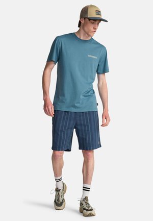 Młody mężczyzna ubrany w niebieską koszulkę z krótkim rękawem, granatowe szorty w paski, beżowe trampki, białe skarpetki z czarnymi paskami oraz beżowo-niebieską czapkę, idzie.