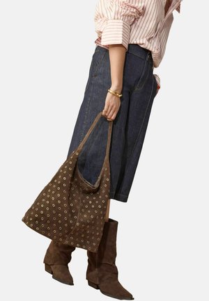 Frau trägt weite Jeanshosen, gestreiftes Hemd, braune Wildlederstiefel und hält eine braune Wildledertasche mit goldenen Ösensdetails neben sich.