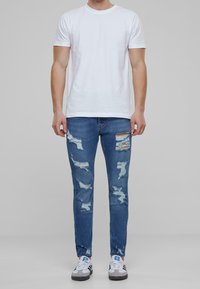 Camiseta blanca de cuello redondo, jeans skinny azules desgastados con detalles rasgados, y zapatillas gris con acentos negros y logo azul.