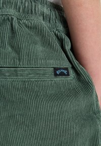 Groene corduroybroek met een gestructureerd oppervlak, voorzien van een elastische tailleband en een klein zwart label met blauwe tekst.