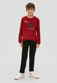 Czerwona bluza z nadrukiem tekstowym, czarne spodnie i białe sneakersy. Bluza ma długie rękawy i okrągły dekolt, gładką teksturę.