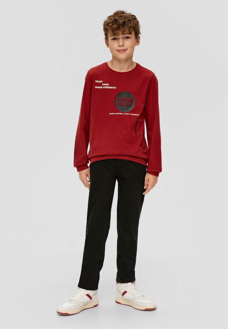 Czerwona bluza z nadrukiem tekstowym, czarne spodnie i białe sneakersy. Bluza ma długie rękawy i okrągły dekolt, gładką teksturę.