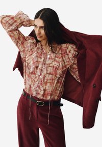 Světlounce růžová halenka s volánky a károvaným vzorem, doplněná tmavě červenými manšestrovými kalhotami a odpovídajícím sakem. Černý pásek se zlatou přezkou jako akcent.