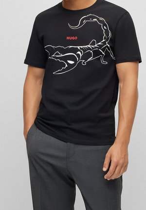 Person wearing black t-shirt with large white scorpion graphic and red "HUGO" text, paired with gray pants, hands in pockets.