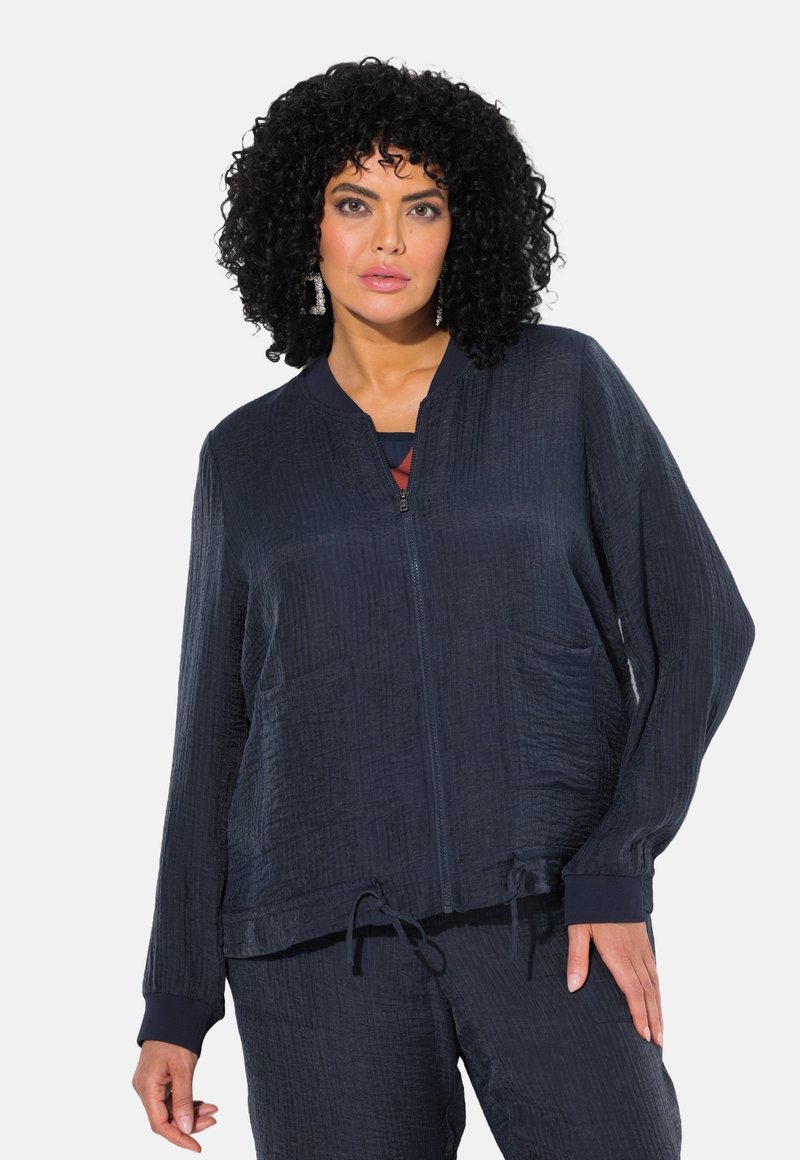 Giacca zip-up blu navy con texture stropicciata, vestibilità comoda e tasche laterali. Presenta un collo senza colletto e polsini elasticizzati.
