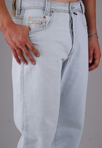 Hellblaue Jeans aus Jeansstoff mit Knopfleiste, getragen von einer Person mit einer Narbe an der linken Hand und einem silbernen Armband am rechten Handgelenk.