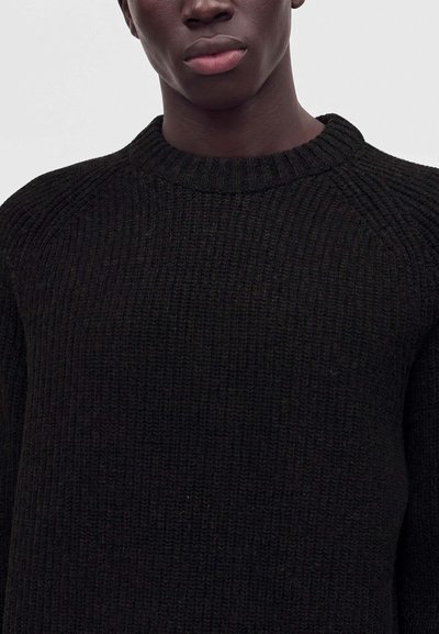 Gros plan d'une personne portant un pull en maille côtelée noire avec un col rond sur un fond clair uni.