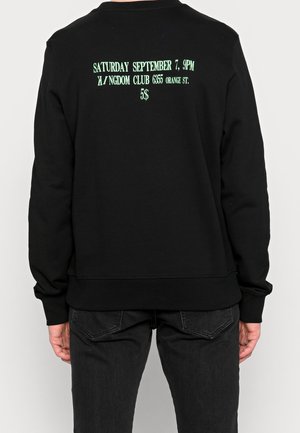 Sudadera negra que lleva puesta una persona, el texto en la parte de atrás dice: "Sábado 7 de septiembre, 9PM Kingdom Club 6355 Orange St. 5$" en fuente verde.