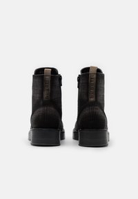 Schwarze Knöchelstiefel mit einem Obermaterial aus strukturiertem Stoff, Reißverschluss und einer flachen, gerippten Sohle. Die hellbraune Zuglasche hinten ist mit einer Markenkennzeichnung versehen.