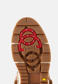 Brązowa gumowa podeszwa buta z jodełkowym bieżnikiem, czerwonymi akcentami i okrągłymi wzorami, z teksturowanymi rowkami oraz logo w kolorach żółtym i białym.