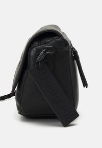 Sac à main en cuir noir texturé avec un rabat, une fermeture éclair latérale, une sangle tressée et une grande étiquette en caoutchouc portant la mention "Desigual".