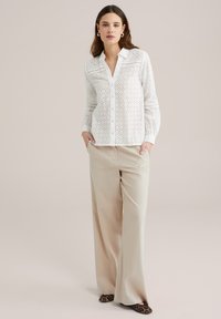 Blusa bianca ricamata con bottoni anteriori, maniche lunghe e colletto a punta; abbinata a pantaloni beige a gamba larga e scarpe con stampa leopardata.