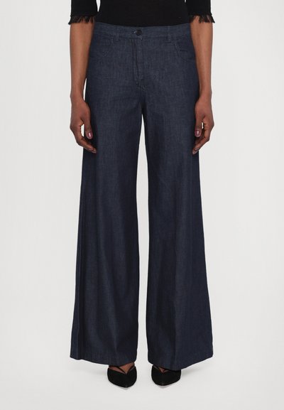 Pantalons en denim foncé à jambes larges avec une fermeture à bouton, poches cousues et une texture lisse, associés à des chaussures noires à bouts pointus.