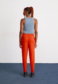 Vrouw met gevlochten haar in een paardenstaart, die oorbellen in de vorm van een ring draagt, een grijs mouwloos topje, een oranje broek en zwarte laarzen aanheeft, staande op een blauwe vloer.