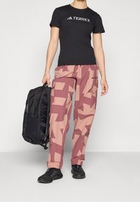 Svart t-shirt med "TERREX" logotyp, mönstrade rosa cargobyxor och svarta sneakers. En svart ryggsäck hålls i vänster hand.