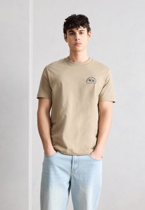 Mladý muž s kučeravými vlasmi nosí béžové tričko s krátkym rukávom a malým tmavým logom, svetlomodré rifle, stojí s rukami v vreckách na jednoduchom pozadí.