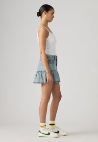 Φούστα από ανοιχτό τζιν με βολάν και ακατέργαστο τελείωμα, συνδυασμένη με λευκή εφαρμοστή φανέλα και λευκά παπούτσια με πράσινες λεπτομέρειες και ριγέ κάλτσες.