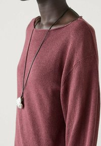 Maglione lavorato a maglia color bordeaux con scollatura ampia, tessuto textured e una collana nera con cordino che presenta un ciondolo argentato.