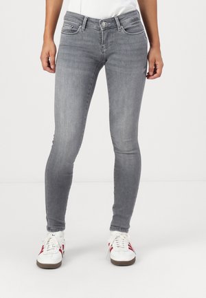 Femme portant un jean gris ajusté et des baskets blanches avec des bandes rouges, debout sur un fond blanc uni.