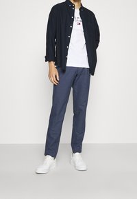 Camisa de botones azul marino sobre una camiseta blanca con logo, combinada con pantalones azules y zapatillas blancas, exhibida en un modelo frente a un fondo neutral.