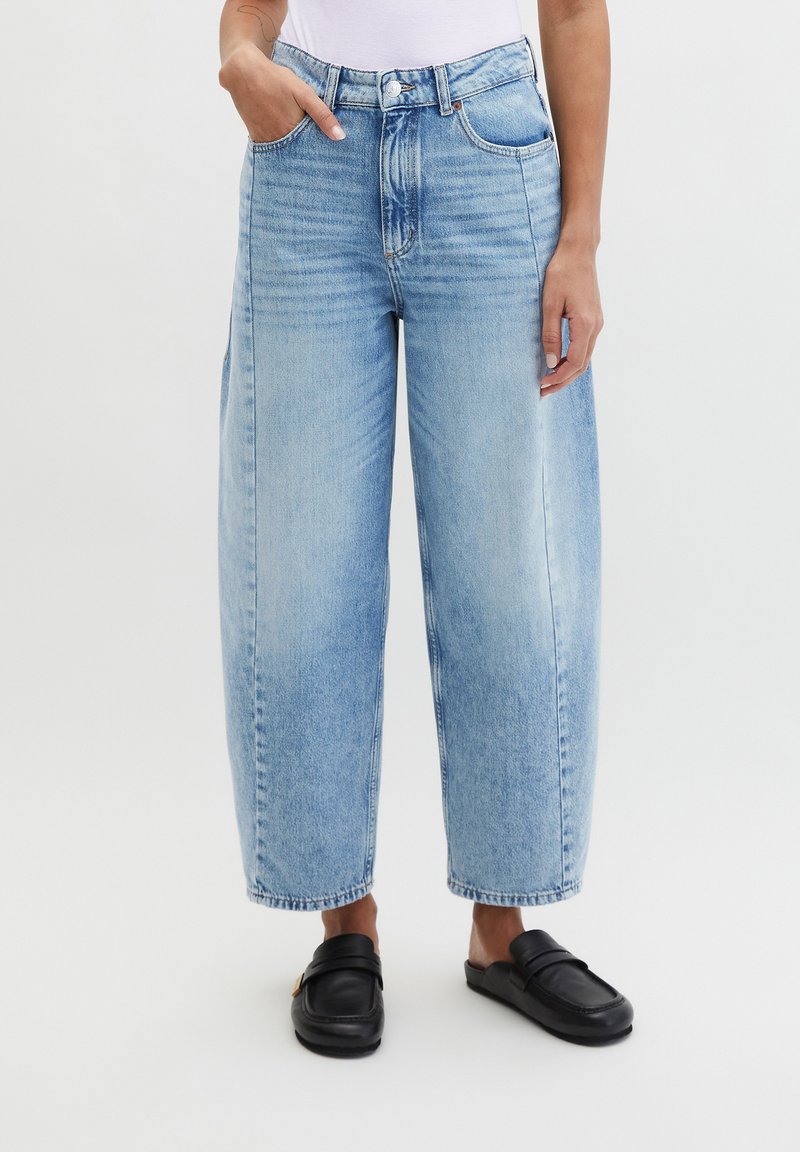 Persoon gekleed in lichtblauwe wijde cropped jeans met voorzakken en zwarte instaploafers, staand tegen een effen achtergrond.